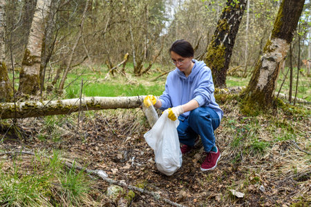 Woman in casual wear, gloves, with plastic trash bag collecting garbage in forest. Plogging