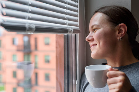 Happy smiling woman with cup looking to window with blinds