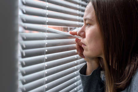 Anxious woman looking to window through blinds and waitingの写真素材