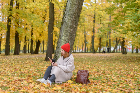 Woman drinking coffee and using phone sitting near tree at autumn park