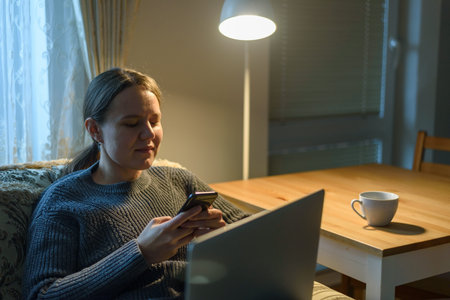 Woman in sweater using her phone sitting near laptop at home living room