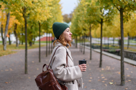 Woman in wool coat and green knitted hat walking and drinking coffee from reusable cup at autumn parkの写真素材