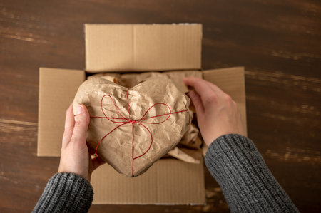 Hands holding Heart-shaped gift box wrapped in kraft paper on wooden background. Just unpacked parcelの写真素材