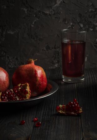 Two pomegranates lie on a metal plate, which stands on a dark wooden table. About them is part of the third broken pomegranate. Behind is a glass of pomegranate juice. Low key, vertical orientation.の写真素材