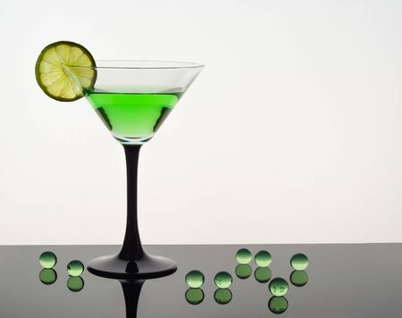 A martini glass with a green drink and a slice of lime on a white background stands on a black glossy surface, next to it are green transparent balls, backlightの写真素材