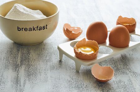 Ingredients for making breakfast on a white wooden table: a cup with the words "breakfast" filled with flour, eggs on a white stand, several broken. Close-up, the lower left corner is free for inscription.の写真素材
