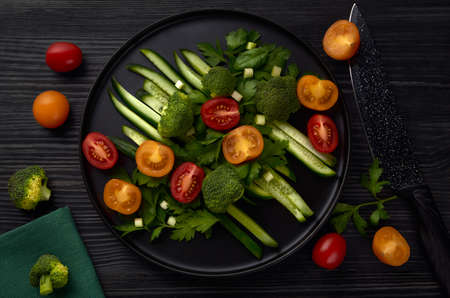 A black round plate of fresh vegetables stands on a black wooden table with a black knife, tomatoes and broccoli nearby. Top view.の写真素材