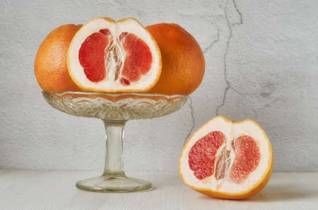 A glass vase with grapefruits stands on a white wooden table against a light wall, one grapefruit is cut, half of it lies on the table.の写真素材