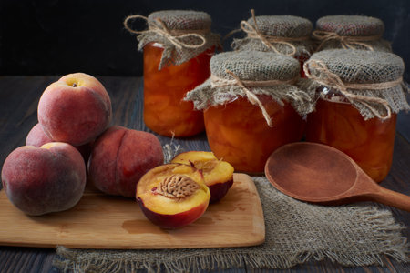 There are several fresh peaches on a cutting board, one of them cut in two. Jars of peach jam sit on a dark wooden table at the back.の写真素材