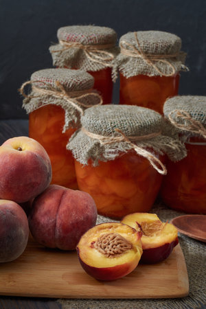 There are several fresh peaches on a cutting board, one of them cut in two. Jars of peach jam sit on a dark wooden table at the back. vertical orientation.の写真素材