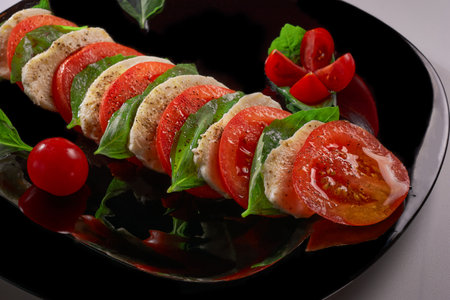 Sliced pieces of tomato, cheese and basil lie on a black plate, close-up.の写真素材