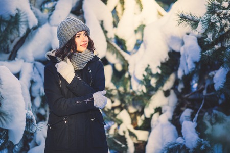 Beautiful young woman in winter jacket, knitted beanie hat and scarf, outdoors in the forest on snowing winter day.の写真素材