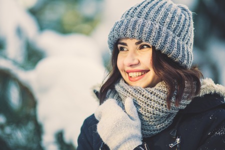 Beautiful young woman in winter jacket, knitted beanie hat and scarf, outdoors in the forest on snowing winter day.の写真素材