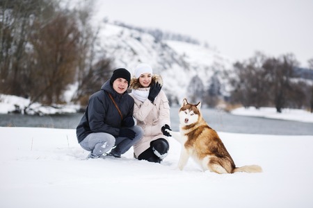 A young pregnant couple walking in the woods with the dog red Husky with blue eyes. Snowy morning in the forest. A girl and a guy dressed in warm clothesの写真素材
