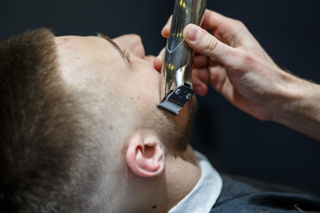 Beard grooming. Side view of young bearded man getting beard haircut by hairdresser while sitting in chair at barbershop.の写真素材