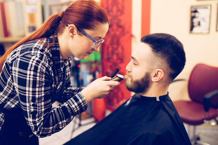 female hairdresser is cutting hair of bearded man client.の写真素材