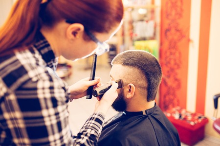 Portrait of handsome young bearded caucasian man getting trendy haircut in modern barber shop. Attractive barber girl working, serving client, doing haircut using metal shears and comb. Indoors shotの写真素材