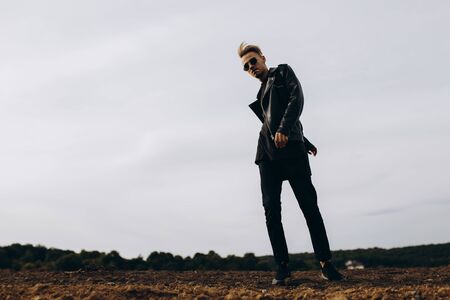 Young sexy man in leather jacket and sunglasses standing outdoor. Silhouette against grey skyの写真素材