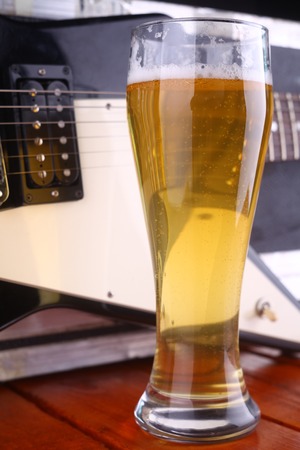 Tall glass full of light beer standing on a wooden table with a music equipment case and guitar in the backgroundの写真素材