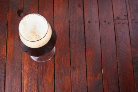 Glass full of dark beer with a rich foam head standing on a wooden tableの写真素材