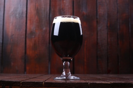 Glass full of dark beer with a rich foam head standing on a wooden tableの写真素材