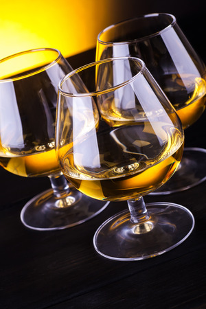 Three snifter glasses with brandy standing on a wood table over a yellow lit backgroundの写真素材