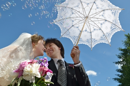 A just married couple, happy bride and groom surrounded soap bubbles, about to kiss in sunshine under umbrella on their wedding dayの写真素材