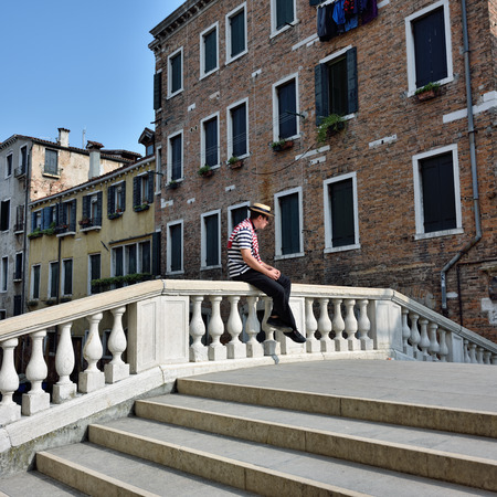 VENICE, ITALY - SEPT 27, 2014: Unidentified man in gondolier dress sits on the bridge fence in Venice. Gondolas trip is the most popular tourist entertainment in Venice,  famous UNESCO World Heritage Siteのeditorial素材