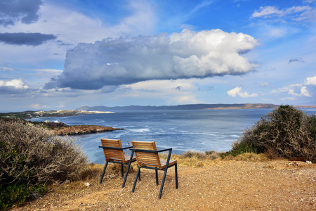 Two empty chairs on high coast of the Cape Sounion on the Aegean sea. View on the coastline and dramatic cloudy sky at sunset time, Attica, Greeceの写真素材