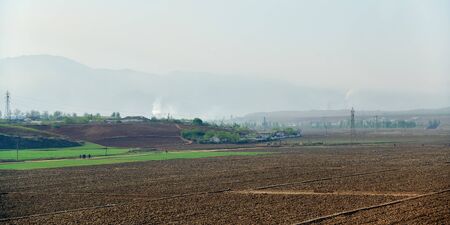 Countryside landscape, North Korea. Village, cultivated agricultural fields, mountain and big chemical plant in backgroundの写真素材