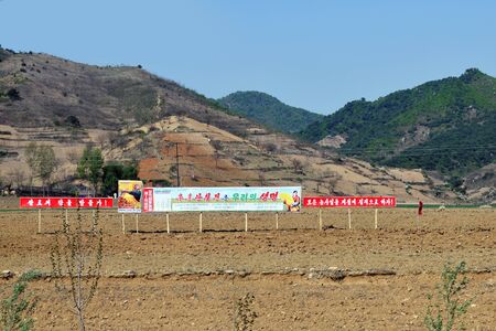 Wonsan region, North Korea - May 5, 2019: Cultivated an agricultural fields. In the foreground is a poster with the results of collective farmers team competition.のeditorial素材
