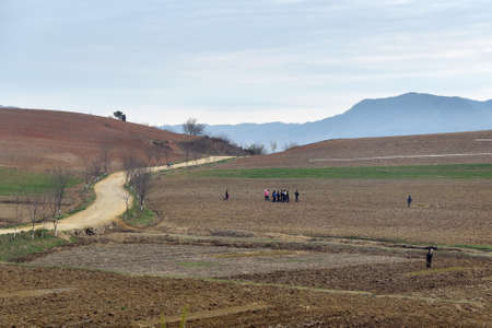 North Korea - April 29, 2019: Hills, dirt road and plowed agriculture fields in foreground. Peasants gathering to work in a fields at dawnのeditorial素材