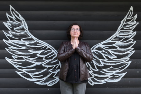 Mature woman in glasses with painted angel's wings stands in a praying pose looking at the sky. Arms folded on chestの写真素材