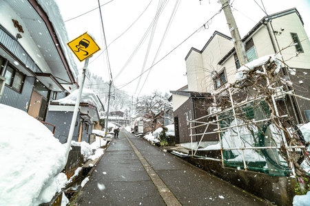 A quiet, narrow street in a Japanese residential area blanketed with fresh snow during winter. A yellow warning sign indicates a slippery road ahead, highlighting the challenging driving and walking conditions in the cold season.の写真素材