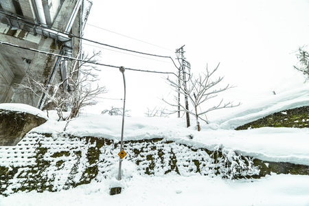 A dramatic scene of a retaining wall covered with moss and topped with thick snowdrifts during a heavy snowfall. The bleak winter atmosphere is emphasized by the bare trees, utility poles, and the presence of a large concrete structure in a cold, remote Japanese setting.の写真素材