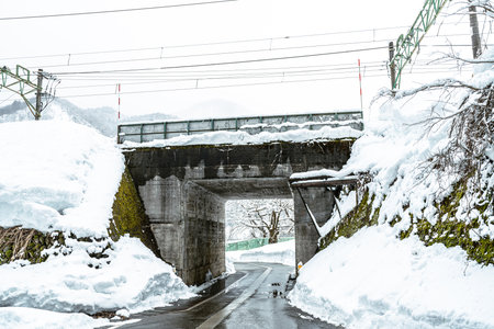 A railway overpass built with concrete and steel crosses a narrow road in a snowy and mountainous area of Japan. Heavy snowdrifts line the road leading through the tunnel-like structure, highlighting the challenging infrastructure and winter travel conditions.の写真素材