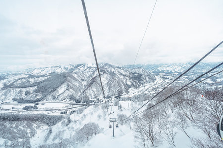 An exhilarating high-angle view from a ski gondola, showing the vast snow-covered mountain landscape, distant valley, and the ski resort slopes below. The cable lines lead the eye into the cold, majestic winter scene.の写真素材
