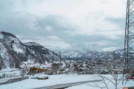 Atmospheric wide view of a remote mountain town or village blanketed in fresh snow on a cold winter day. The valley is surrounded by imposing snow-capped mountains under a dramatic, cloudy grey sky. Captures the stillness and beauty of winter life in a snowy landscape.の写真素材