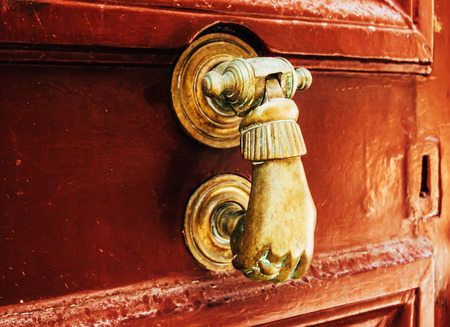 Old bronze door handle in the form of hands on a background of old red scratched wooden door. Street...