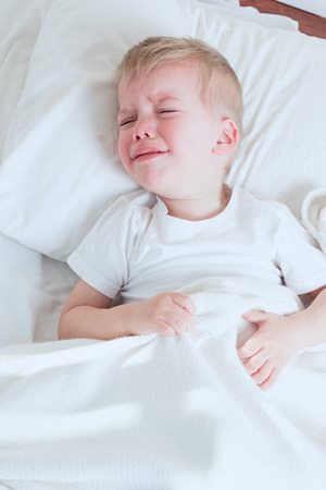 sick toddler boy wearing white tshirt crying in bedの写真素材