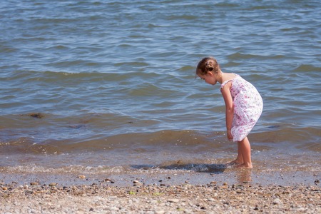 little girl dressed in a long dress looking for shells and rocks in the sea shore on a sunny dayの写真素材
