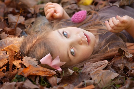 beautiful little girl with brown hair wearing a brown dress laying on the ground covered with leaves in fallの写真素材