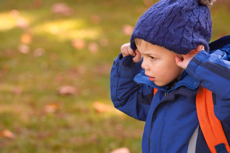little blond boy wearing warm blue hat and blue jacket and a backpack trying to take his hat off outdoorsの写真素材