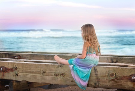 little girl sitting on a bench on a beach and thinking and looking at the oceanの写真素材