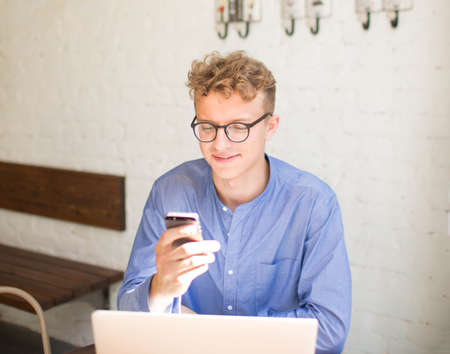 Young man skilled writer reading text message on mobile phone, sitting with laptop computer in...