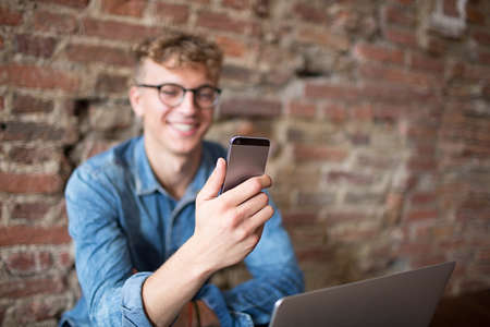 Young smiling hipster guy having video call with friends via mobile phone, sitting against brick...