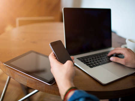 Hipster guy typing text on mobile phone with blank copy space screen, sitting at the table with open...