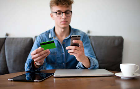 Young man financier booking on-line with credit card and mobile phone, sitting in office interior. Businessman making internet purchase via cell telephone. Shopping store with cellphoneの写真素材