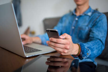 Man skilled employee reading text message on mobile phone, sitting at the table with laptop computer and digital tablet. Hipster guy chatting on modern cell telephone during work on portable net-bookの写真素材