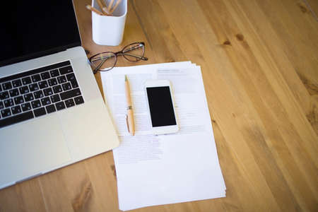 Working space with open laptop computer, modern mobile phone and paper documents. Wooden desktop with portable notebook, cell telephone, contract and tools. Gadget devices on a tableの写真素材